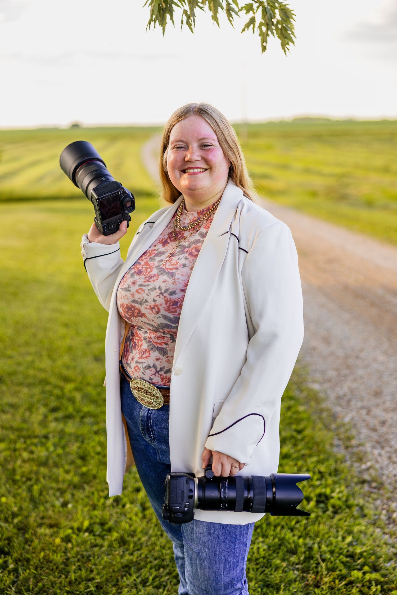 Smiling female photographer holding two cameras outdoors on a rural path surrounded by green fields.