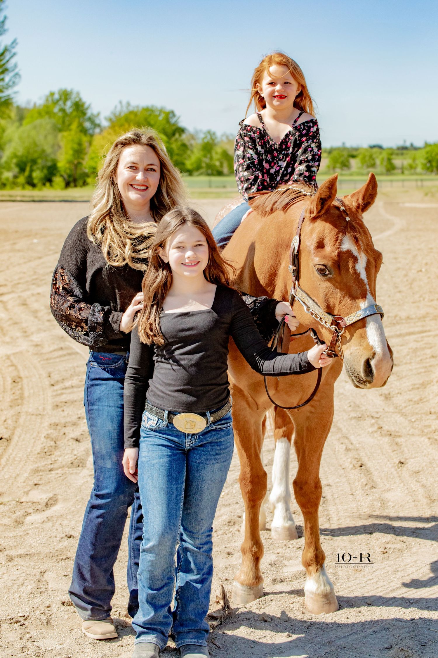 A brown horse stands on a sandy path with riders wearing casual western attire and cowboy boots on a sunny day.