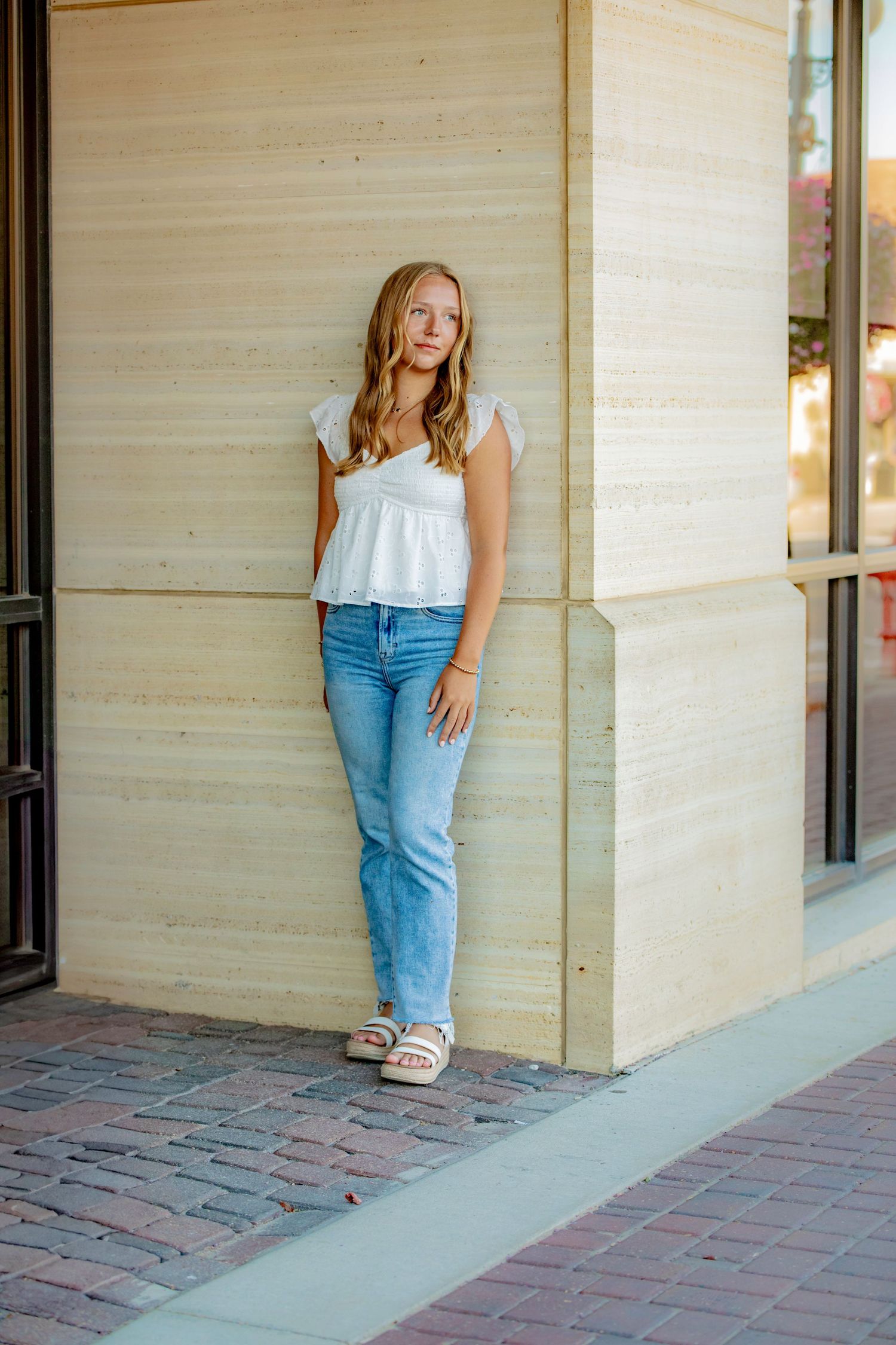 Young person in casual white top and blue jeans poses against beige building wall on brick sidewalk.