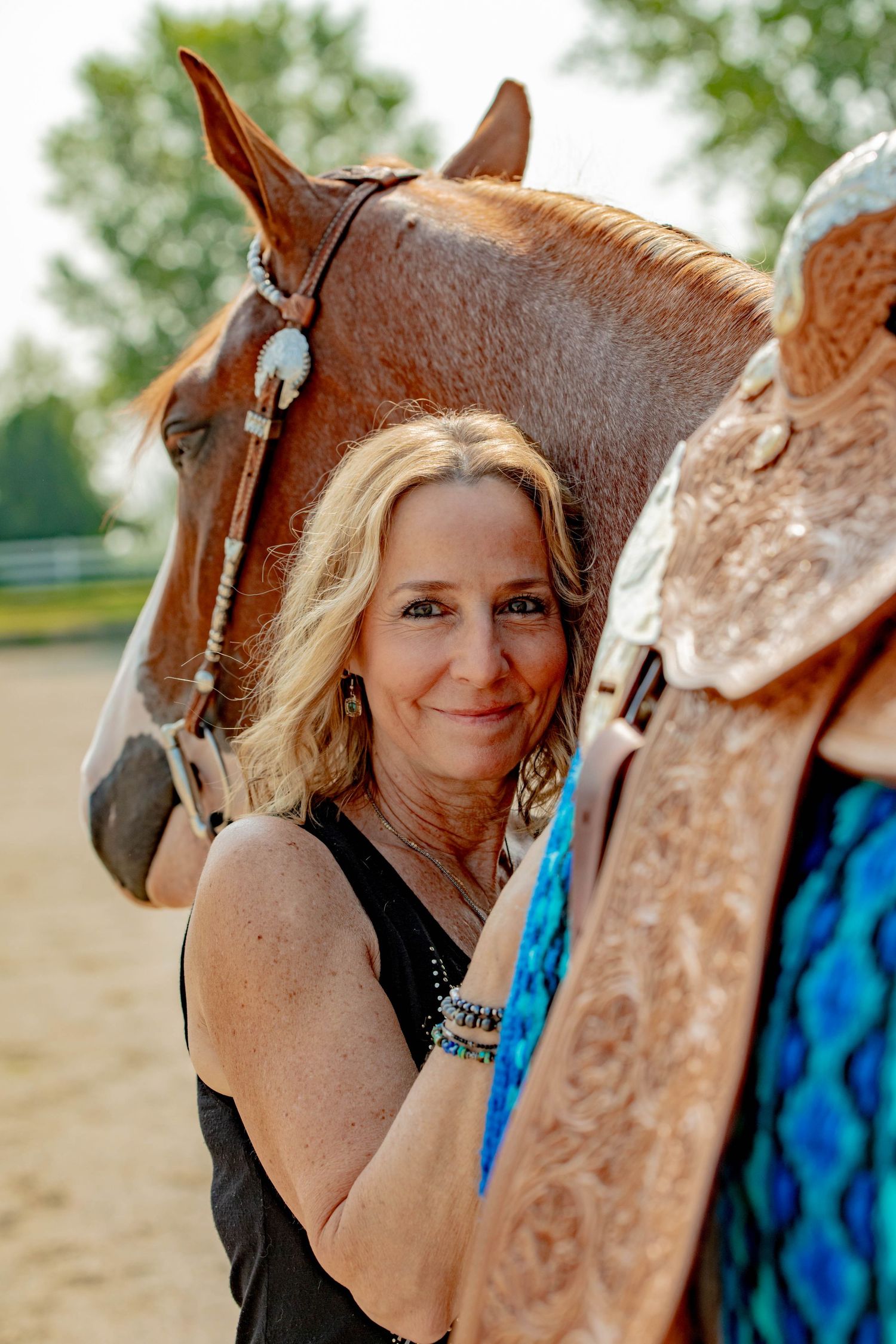 A chestnut horse stands beside a decorative western saddle while being groomed at a ranch on a sunny day.