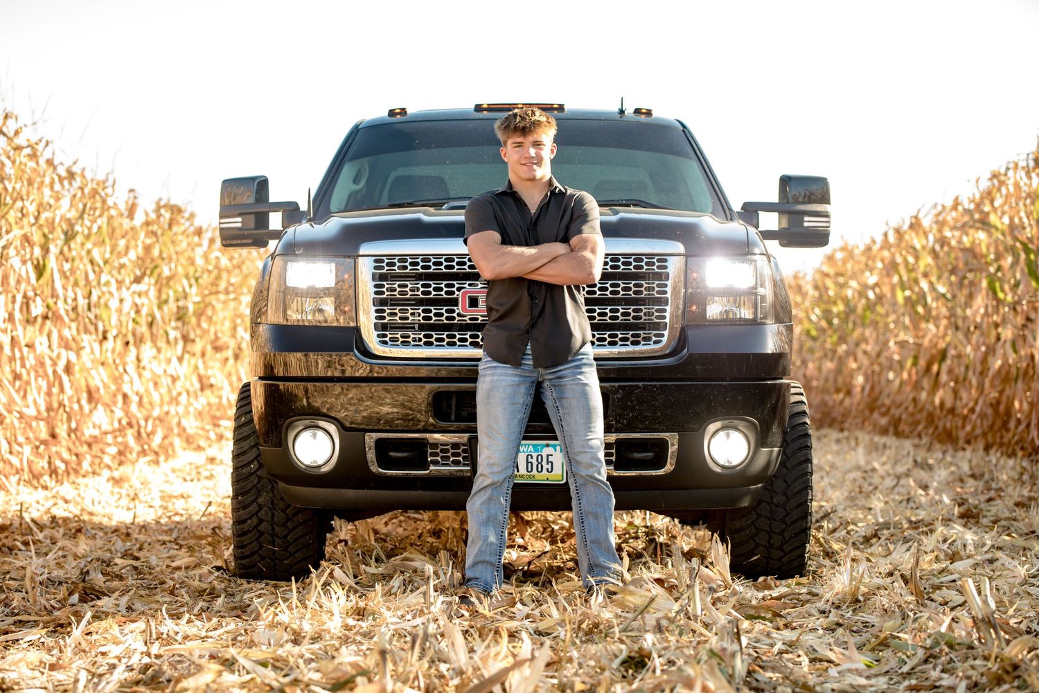 Athletic figure stands with crossed arms in front of large black pickup truck in cornfield.