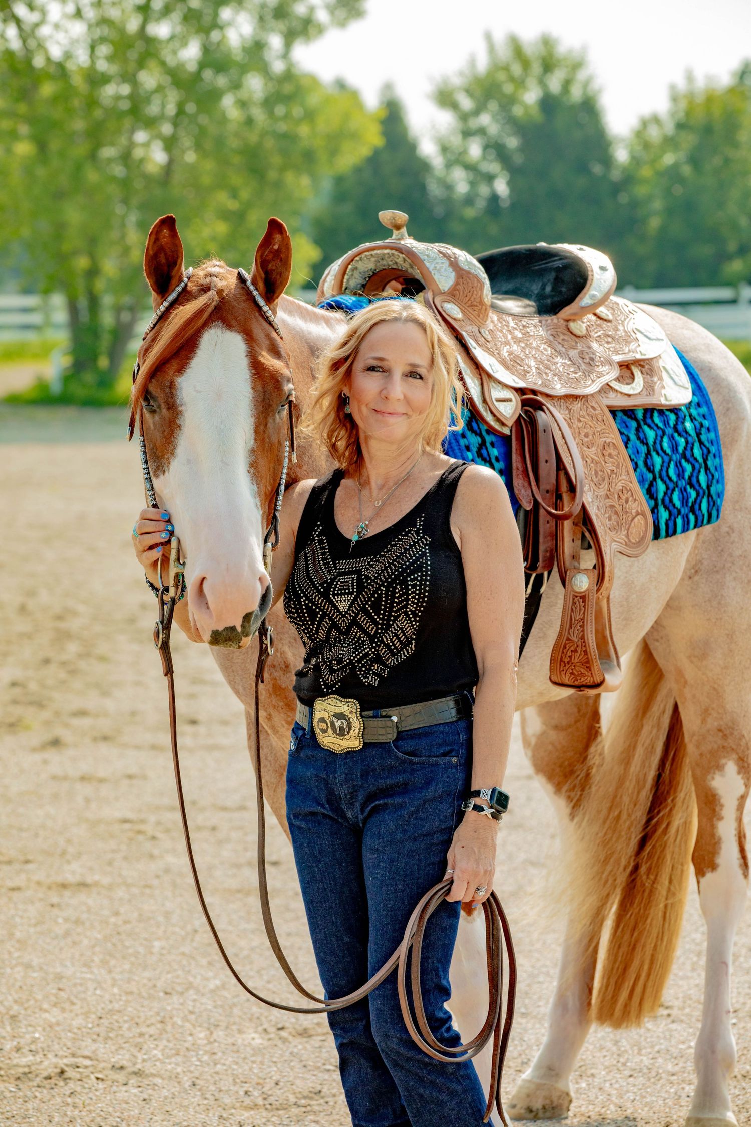 Paint horse with ornate western saddle and blue saddle blanket stands in sunny outdoor arena.
