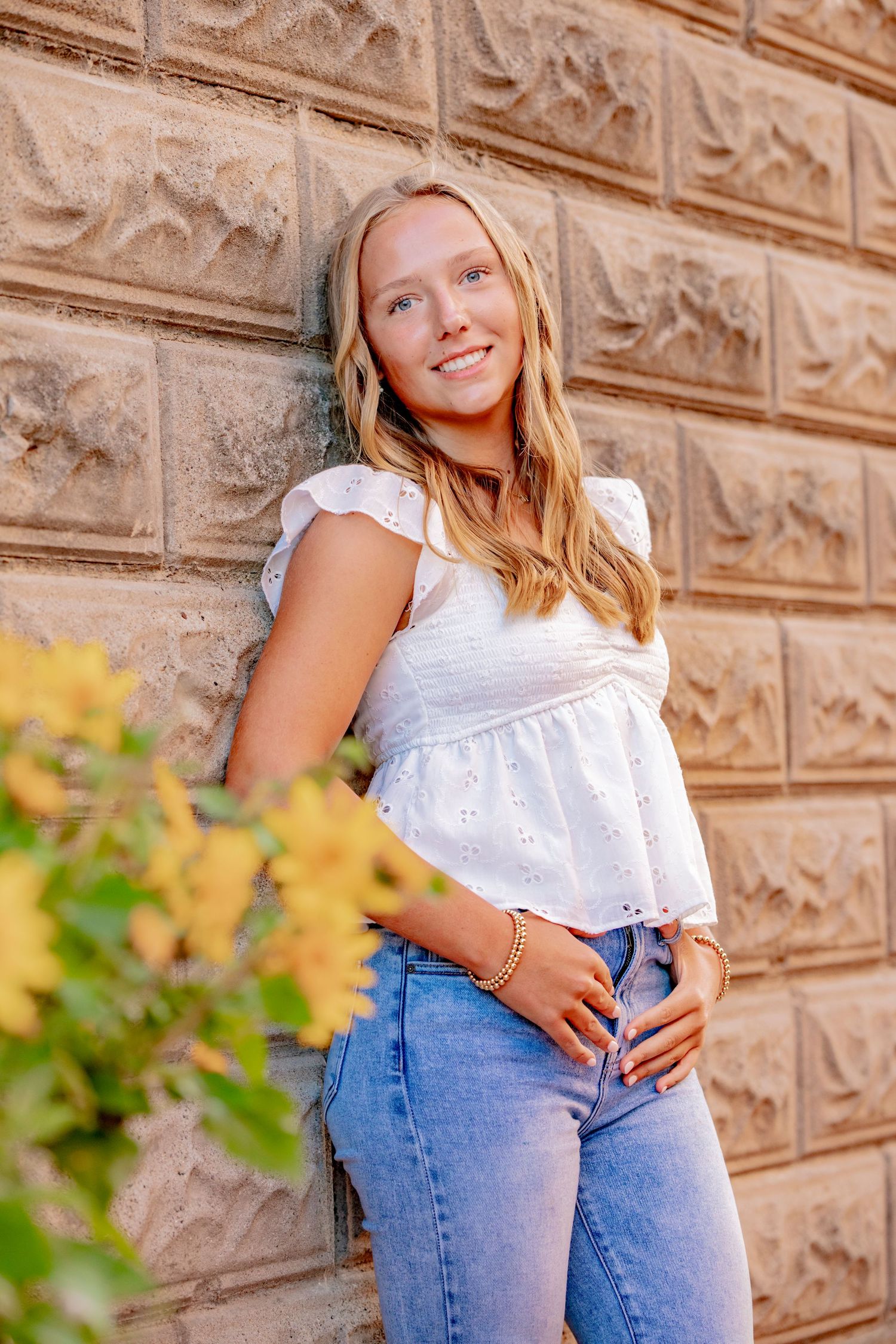 A casual summer outfit with a white eyelet top and blue jeans posed against a textured stone wall.