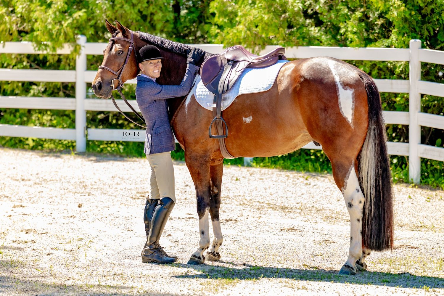Equestrian rider in riding boots and helmet stands beside a chestnut pinto horse with saddle in a white-fenced arena.