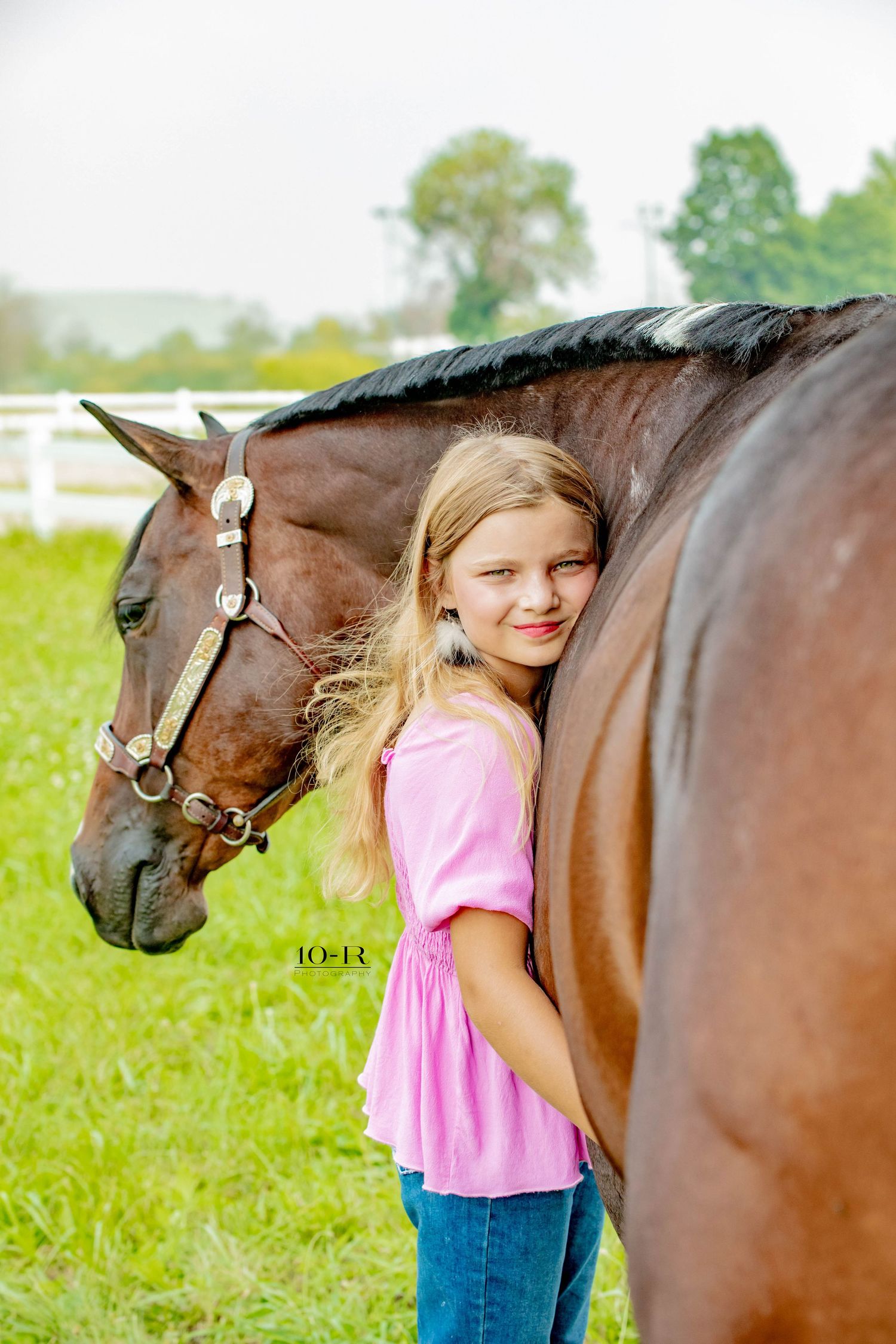 A bay horse standing in a grassy field with jeans and pink shirt in view against a rural fence backdrop.