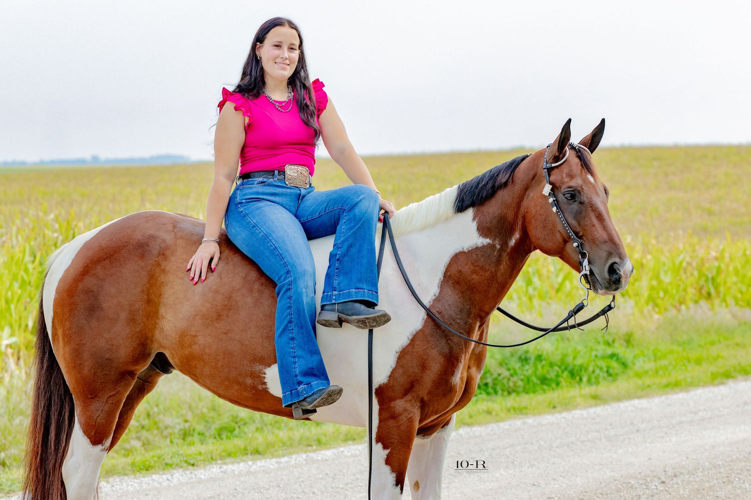 Young woman in pink top and jeans riding a brown and white paint horse on a rural road near cornfields.