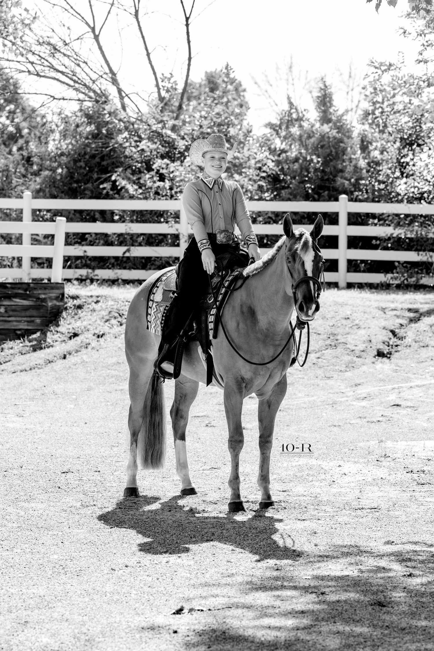 Young rider in western attire sits atop a horse in a fenced arena, black and white equestrian portrait photo.