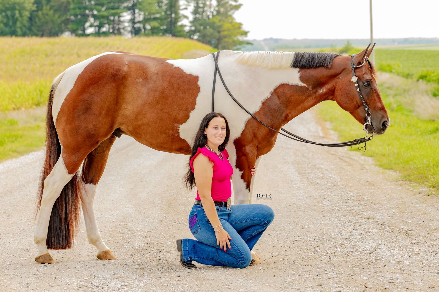 Woman in pink top and jeans kneeling beside a brown and white paint horse on a gravel road.