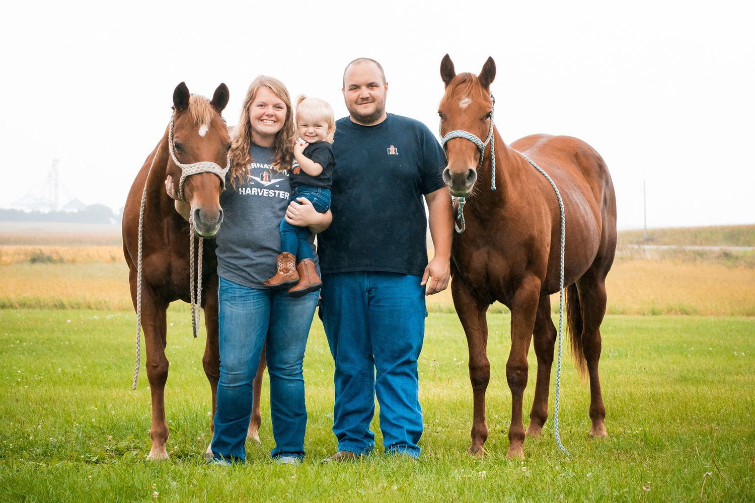 A couple stands in a grassy field with their horses during an outdoor engagement photo session.