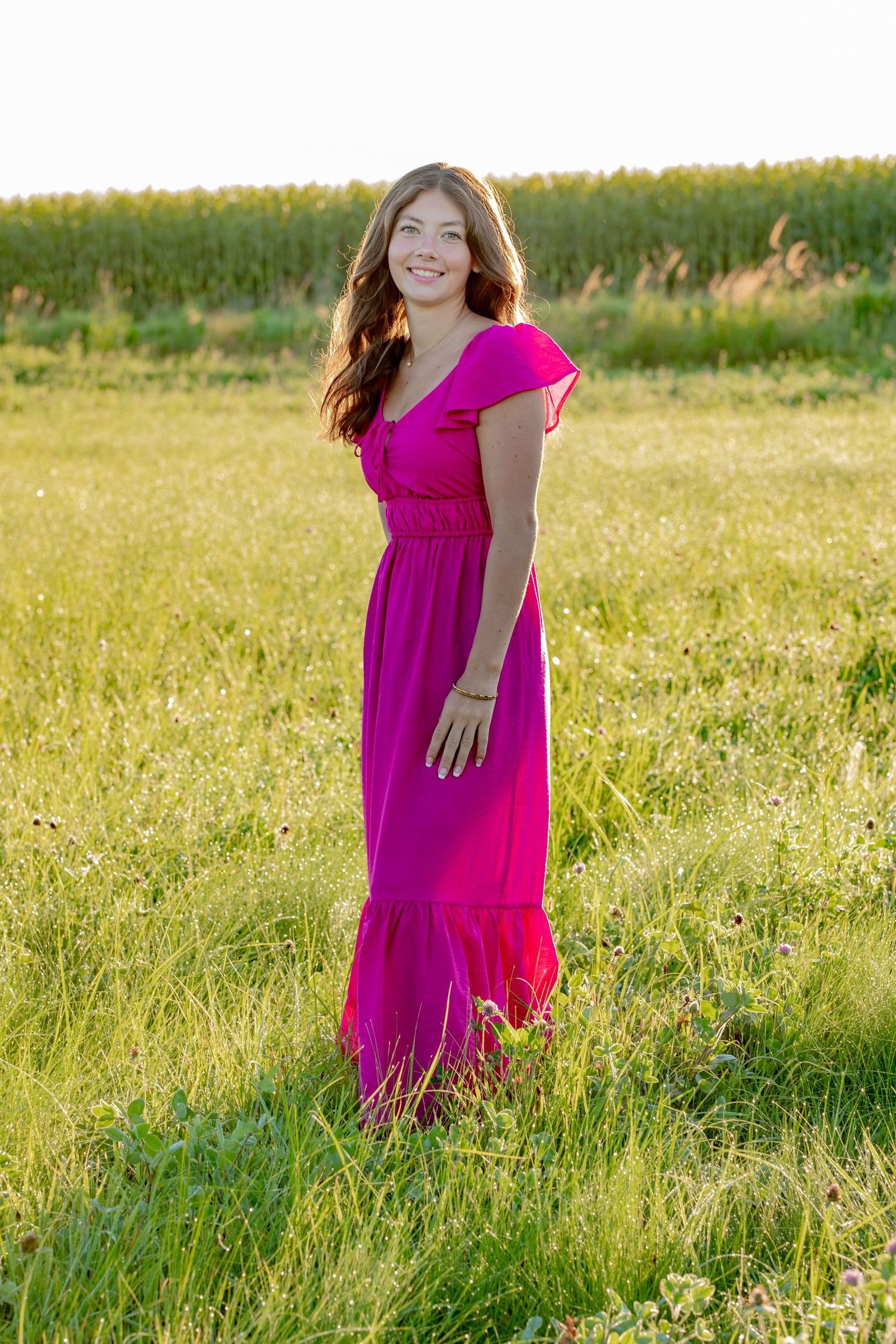 A person in a bright pink dress walks through a sunlit summer meadow filled with green grass and wildflowers.