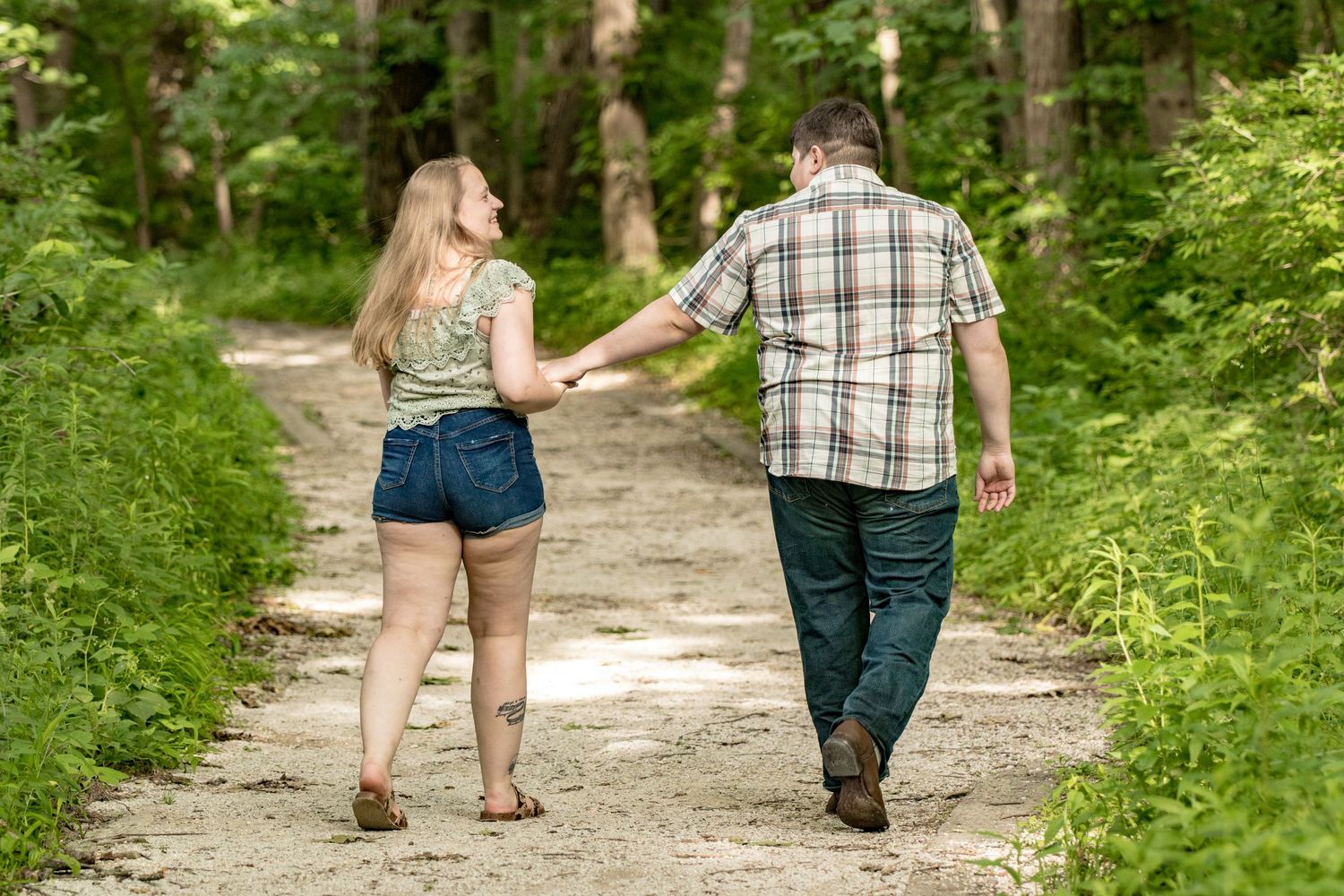 A couple walks hand in hand down a nature trail surrounded by lush green foliage.