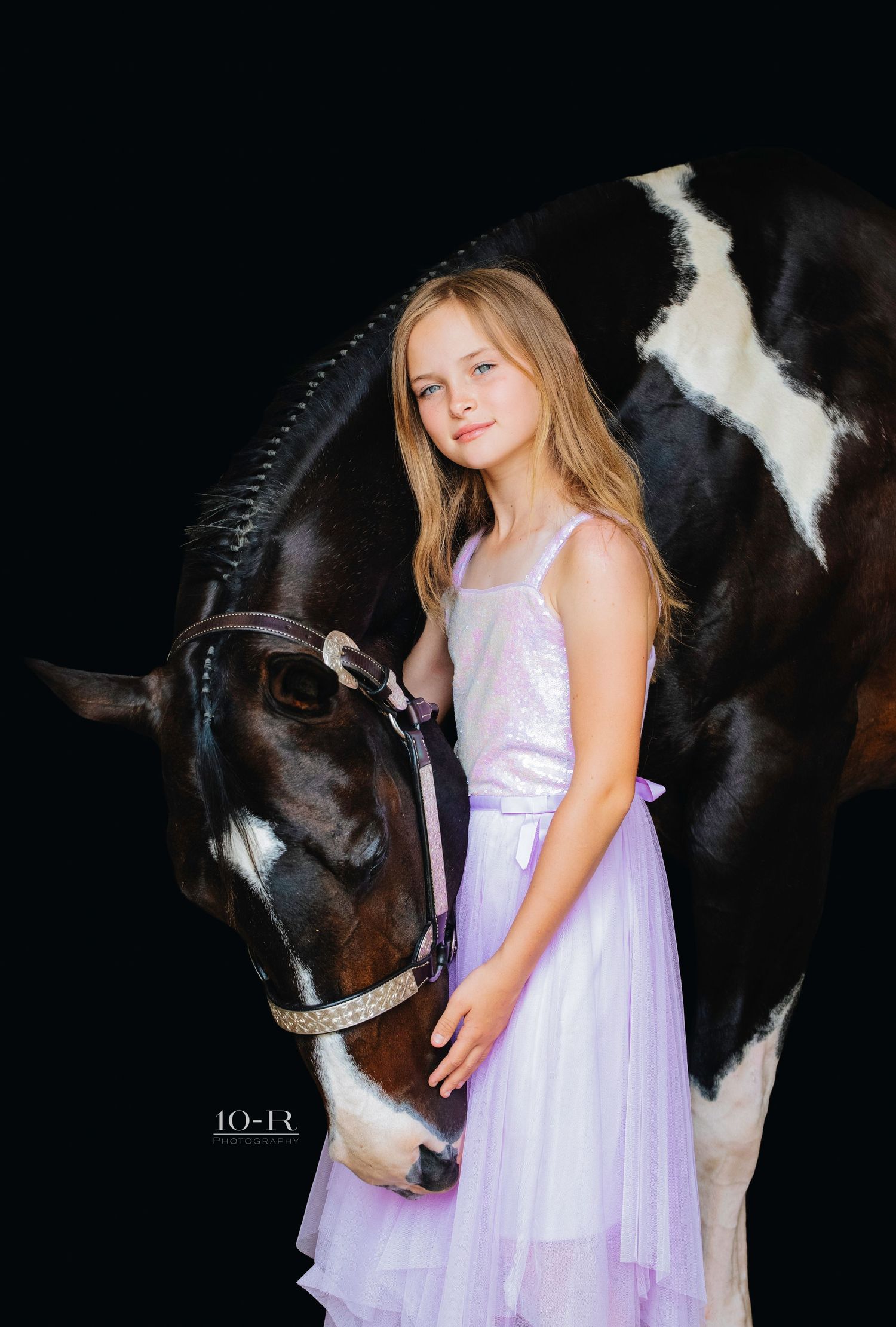 A young girl in a flowing lavender dress poses with a black and white horse in an artistic outdoor portrait series.