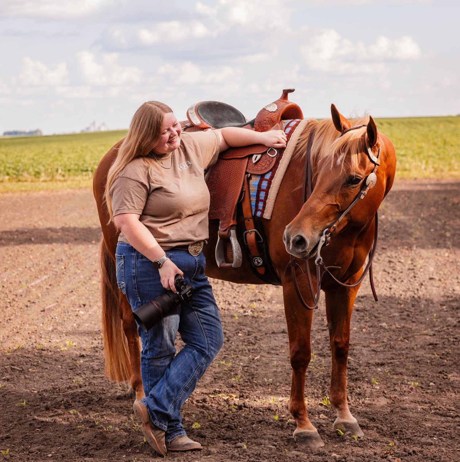 A woman in jeans and tan shirt leans against a saddled chestnut horse on a rural dirt field under a cloudy sky.