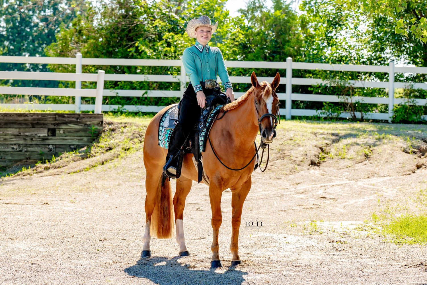 Young rider in turquoise shirt on chestnut horse by white fence in sunny outdoor arena.
