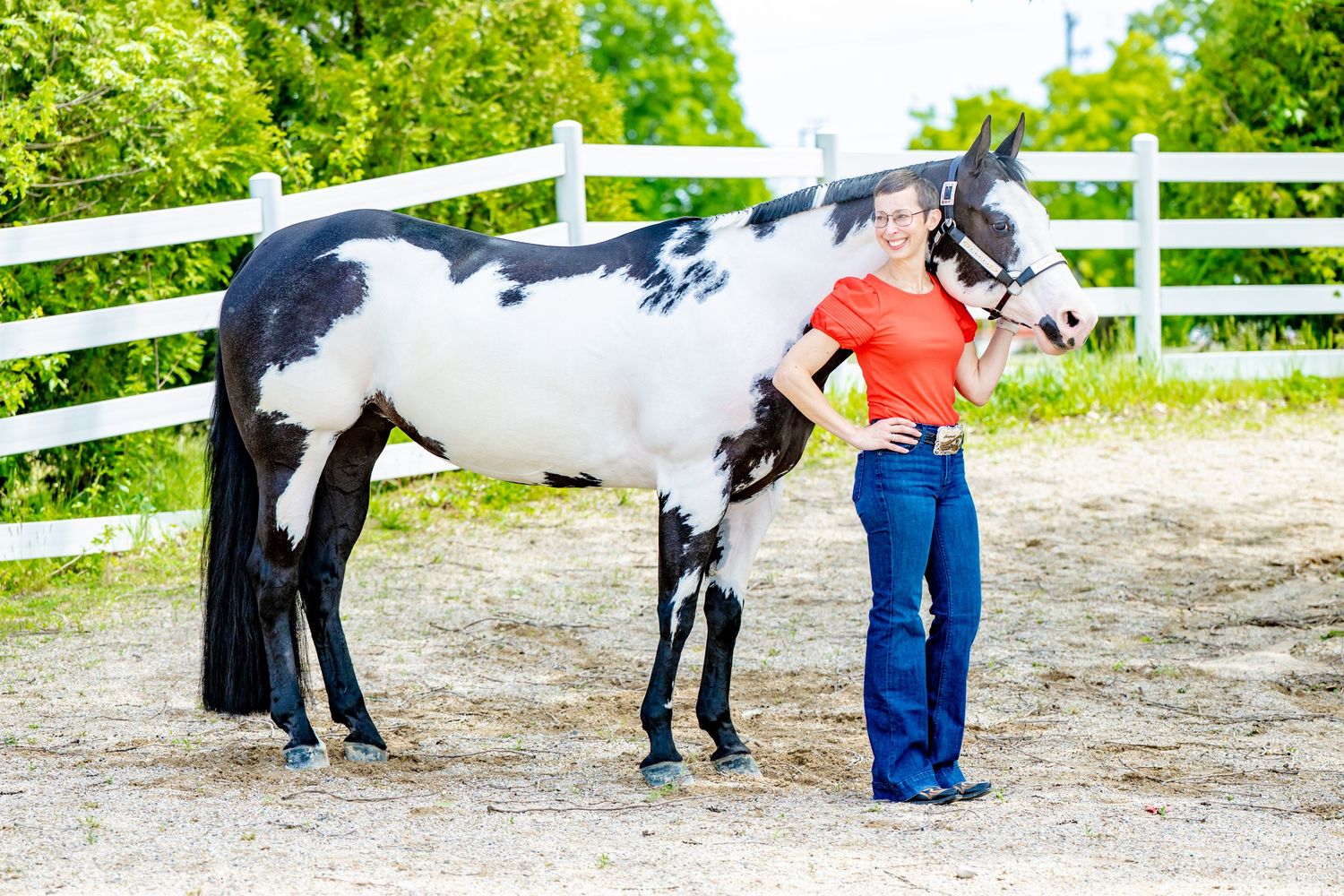 Woman in red top and jeans stands beside a black and white paint horse in a white-fenced arena.