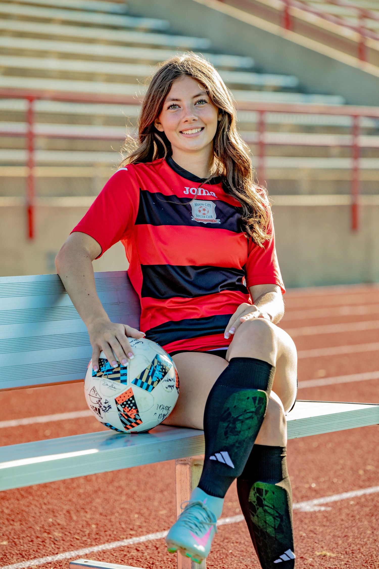 Soccer player in red and black striped jersey sitting on running track with ball.