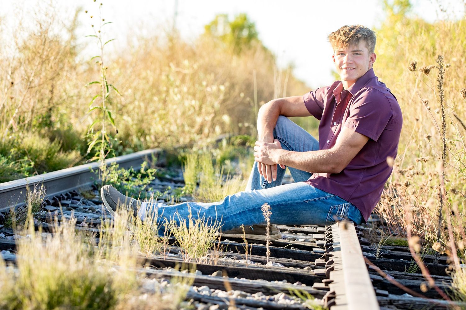 A person in a burgundy shirt sits on railroad tracks surrounded by tall grass at sunset.