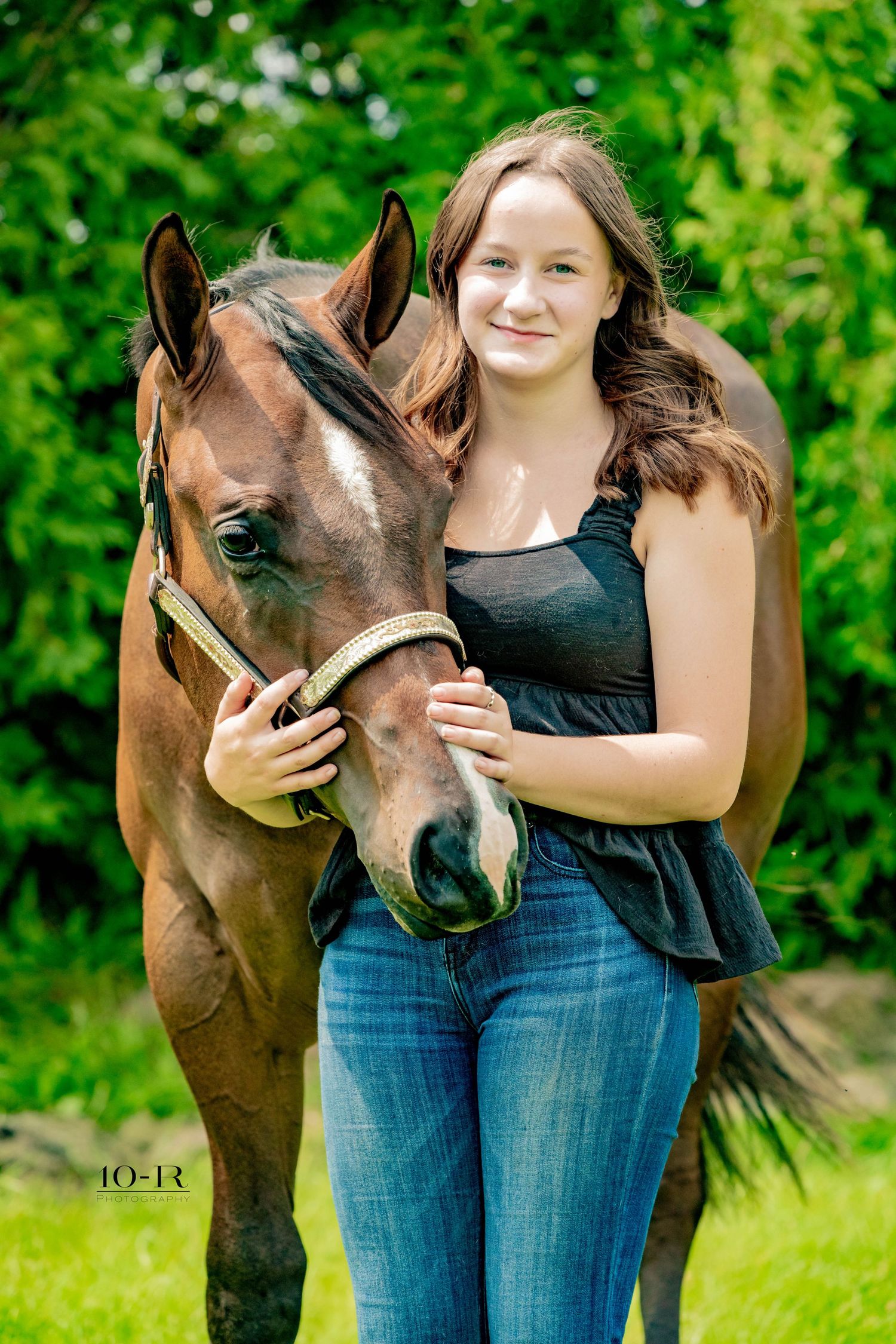 Beautiful brown horse poses with someone on a sunny day against green foliage background.