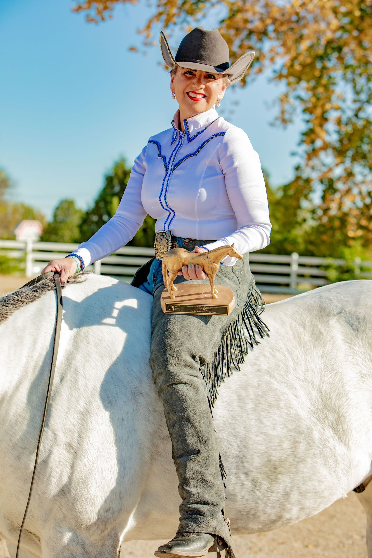 A rider wearing western attire sits atop a white horse against a backdrop of autumn trees and white fencing.