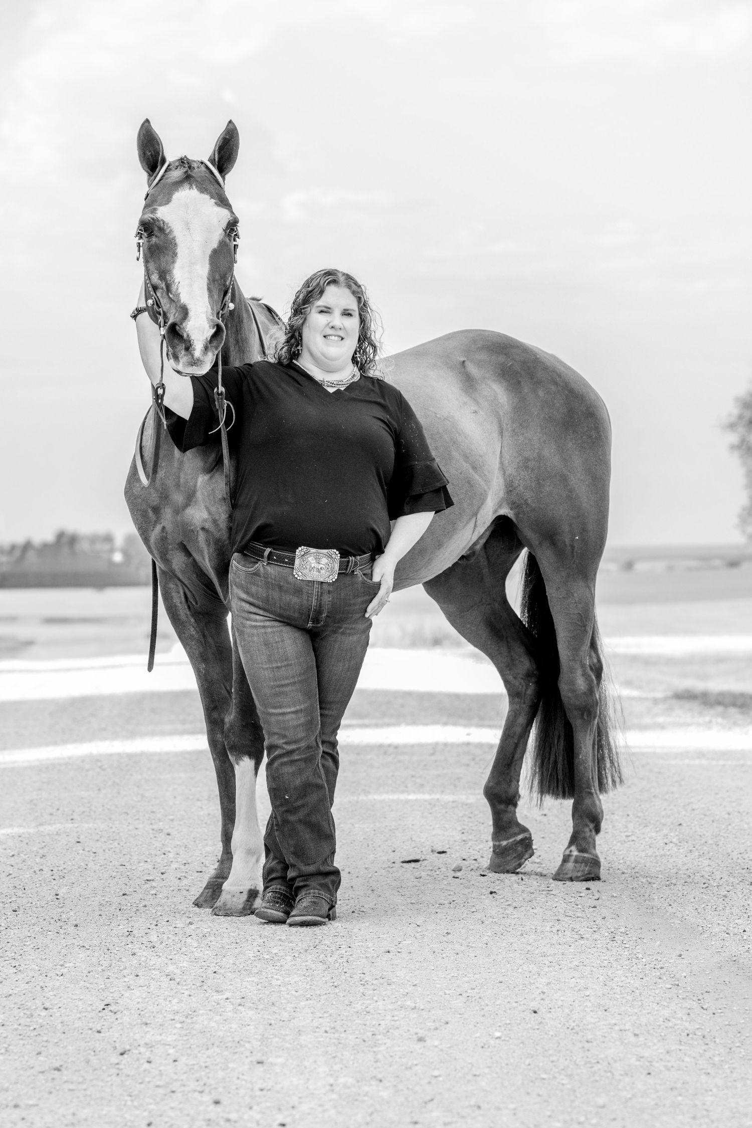 Black and white photo of a woman in jeans and black top standing proudly beside a tall horse on a gravel path.