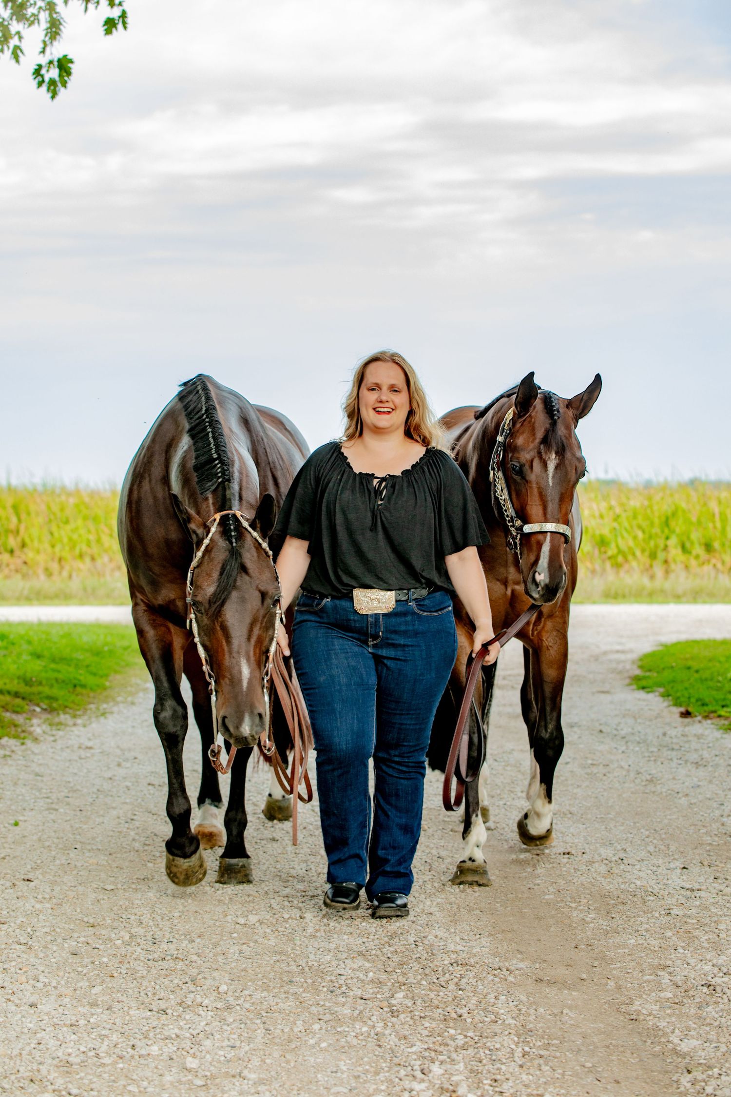Smiling woman walks two chestnut horses on a gravel path with green fields and cloudy sky in background.