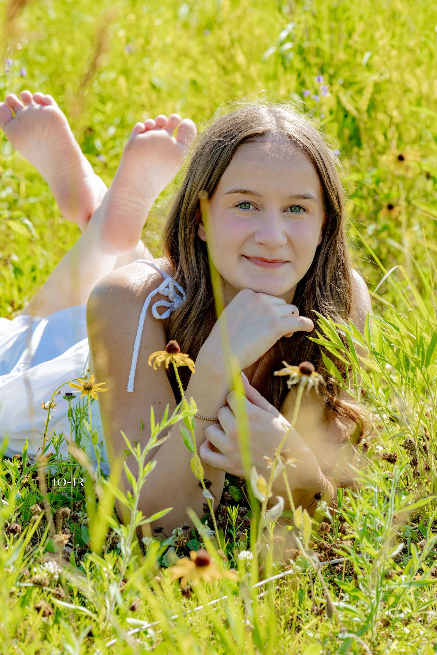 Smiling teenage girl lying in green grass field surrounded by wildflowers on a sunny day.