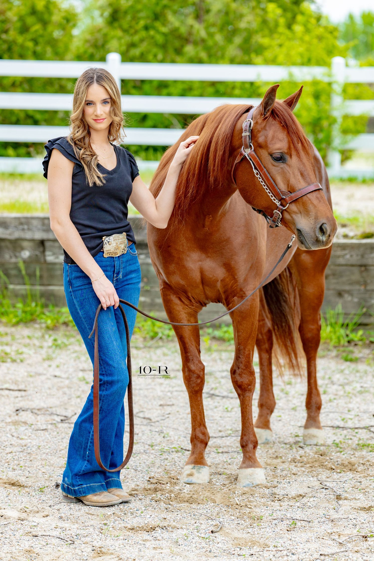 A young woman in jeans and black top stands beside a chestnut horse at a white-fenced ranch.