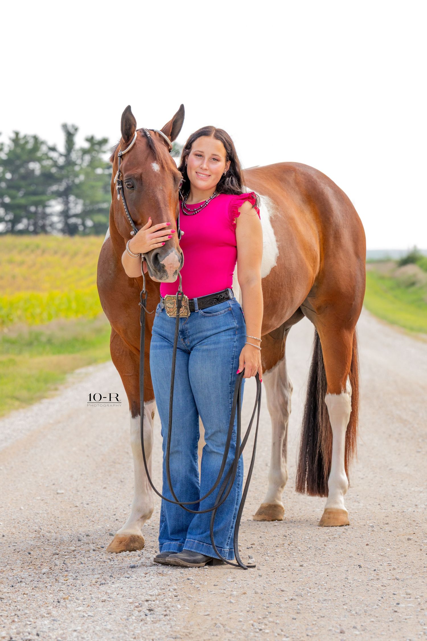 A young woman in a pink top and jeans stands beside a brown and white paint horse on a rural gravel road.