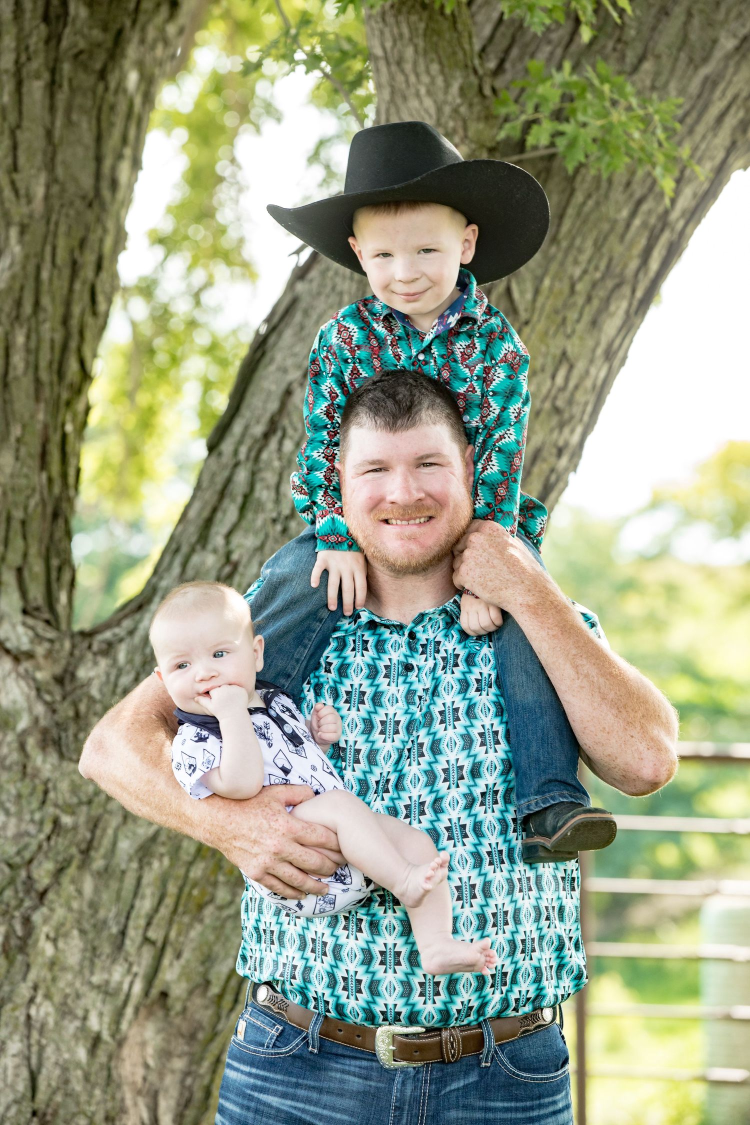 Father carrying two children while posing under a large tree.