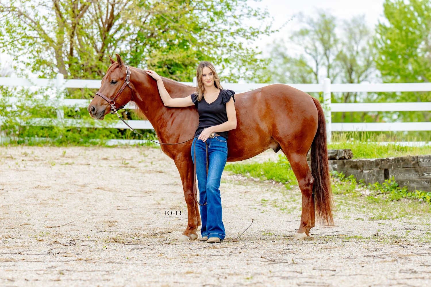 Young woman in jeans and black top posing with a chestnut horse in a sandy paddock with white fencing.