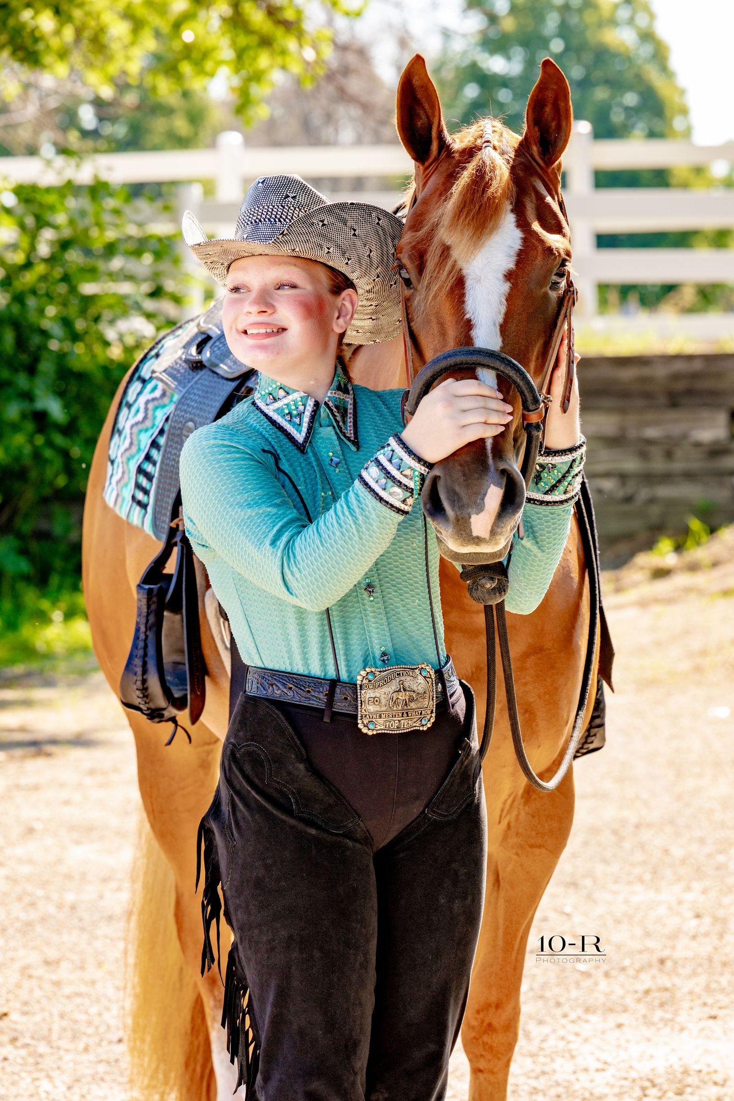 Young cowgirl in turquoise shirt and cowboy hat stands proudly beside a chestnut horse with a blaze marking.