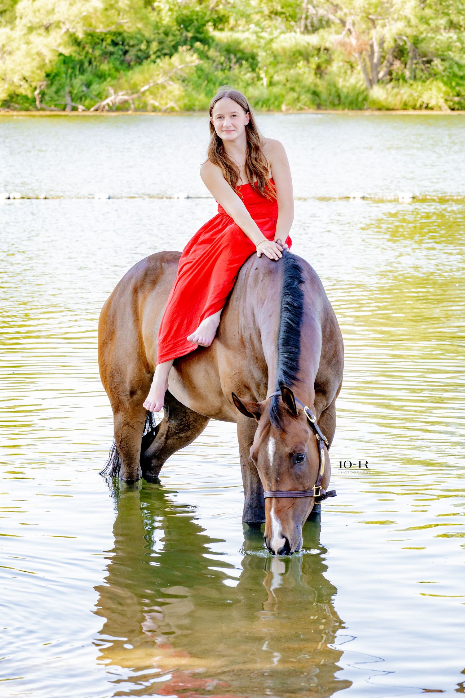 Girl in red dress riding bay horse drinking water in a river.
