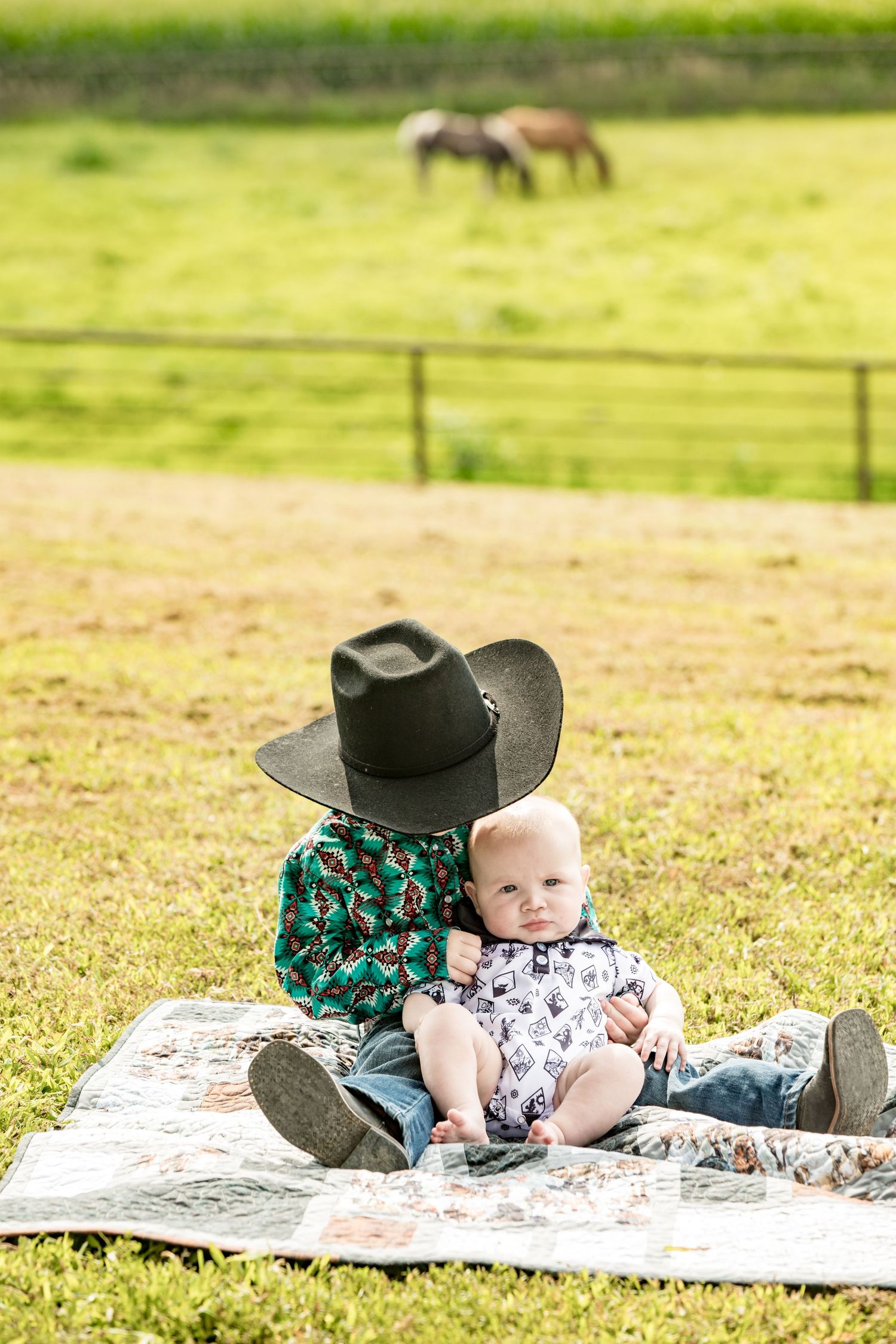Country scene with kids sitting on blanket wearing western attire with horses grazing in background.
