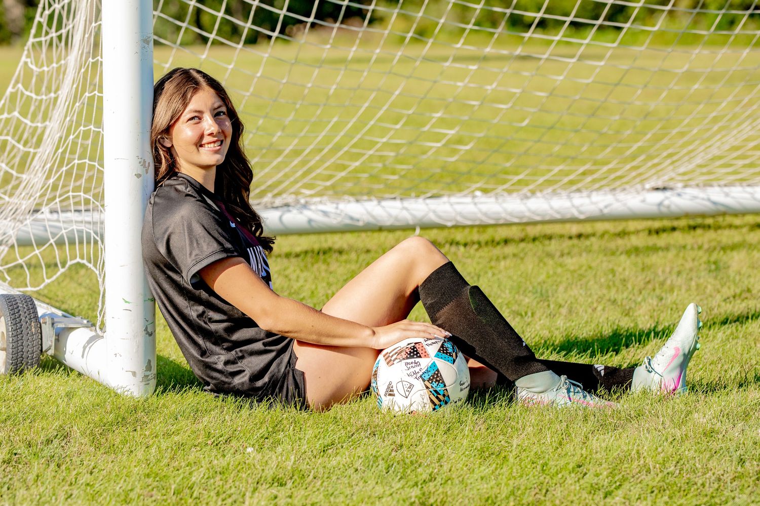 Soccer player relaxing by goal net on sunny field with soccer ball.