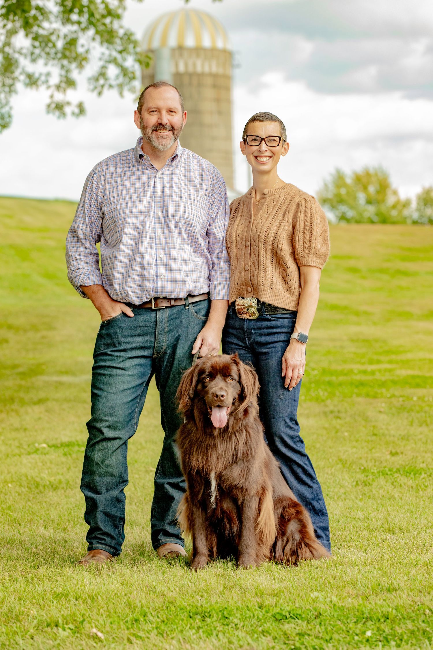 A couple poses with their brown Newfoundland dog on a grassy hillside with farm silos in the background.