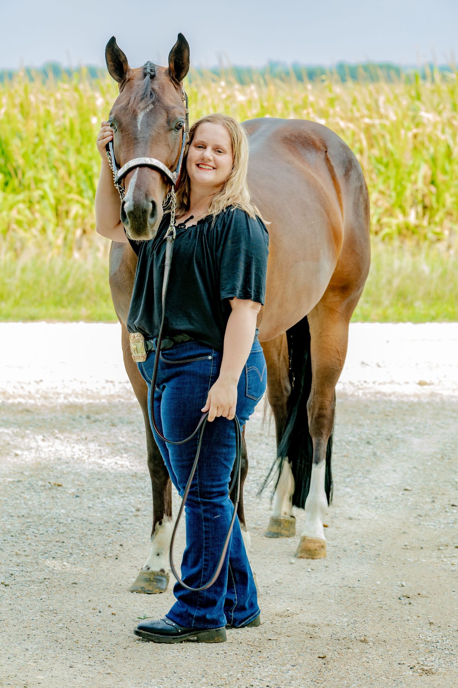 Young woman in jeans and black top smiling beside a brown horse on a gravel path with green fields behind.