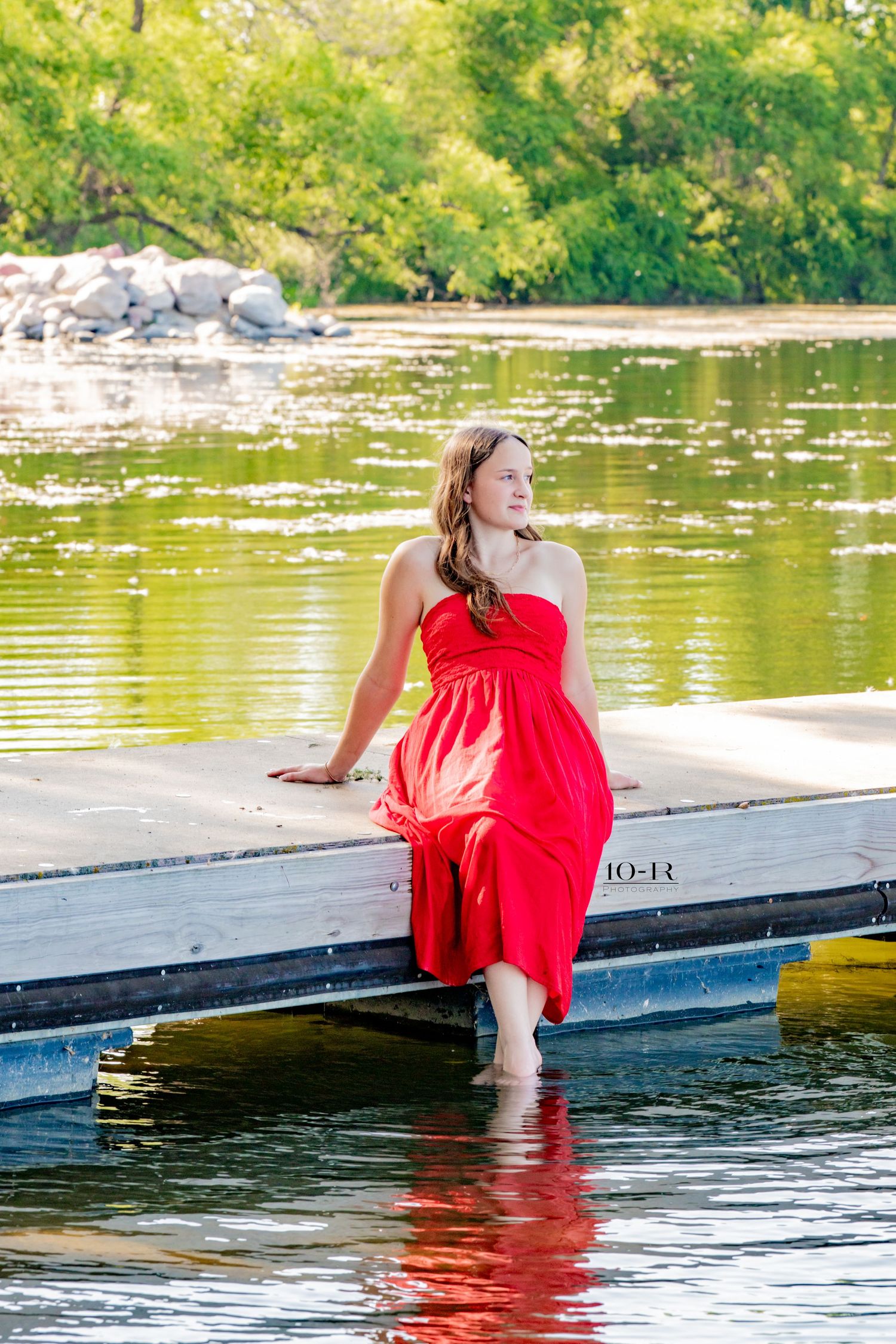 Young woman in flowing red dress sitting on a dock with feet in calm lake water.