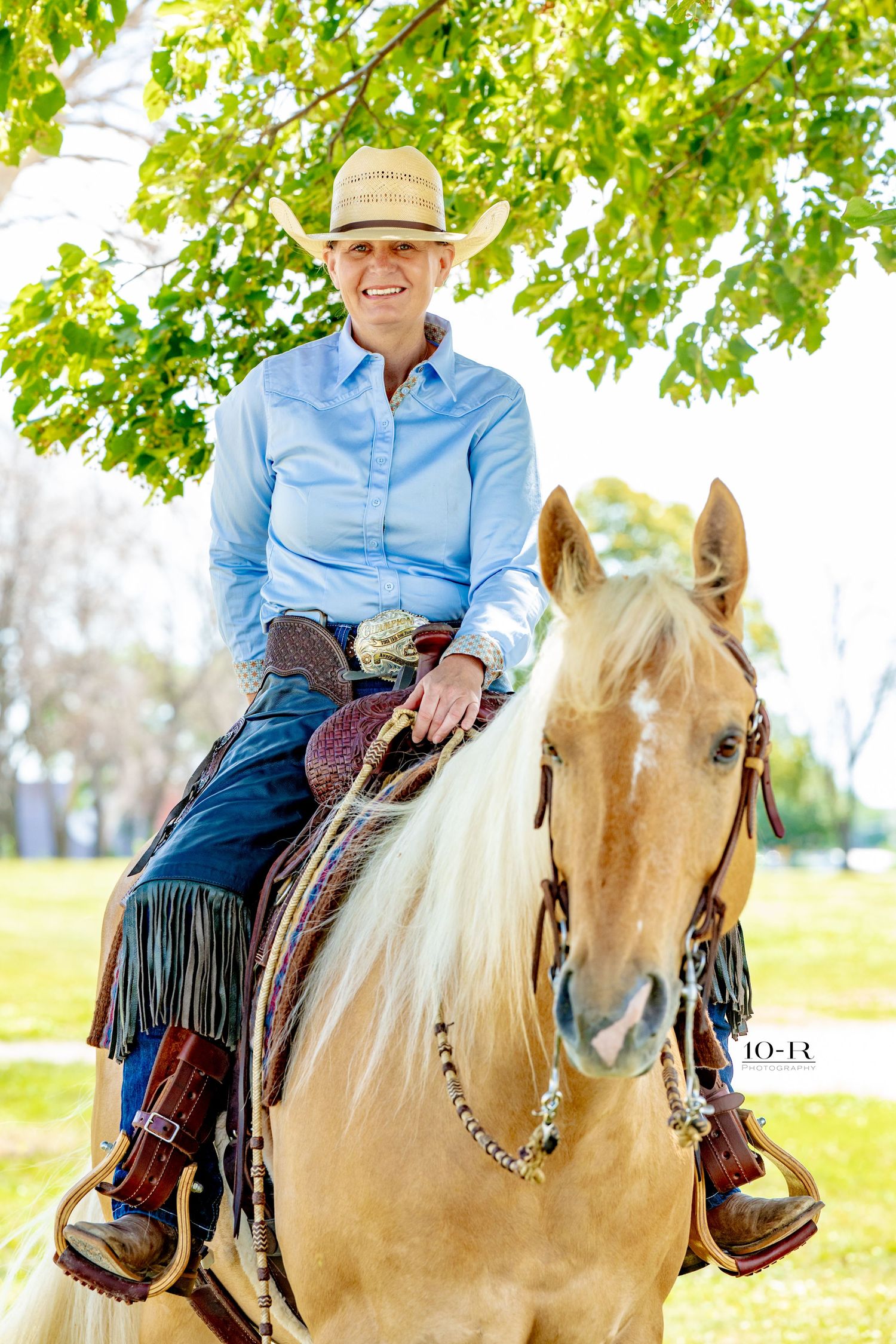 Smiling cowgirl in straw hat and blue shirt riding a palomino horse with white mane under green trees.