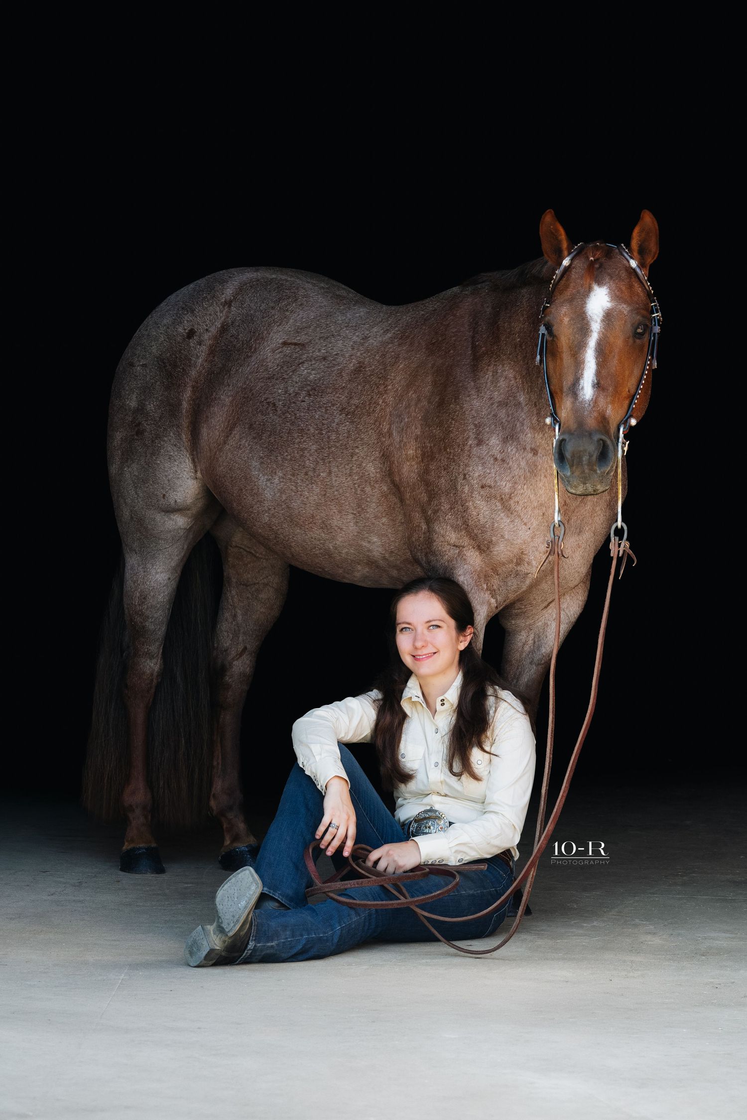 Young woman sitting on ground smiling beside a brown horse with a white blaze, holding reins on dark background.