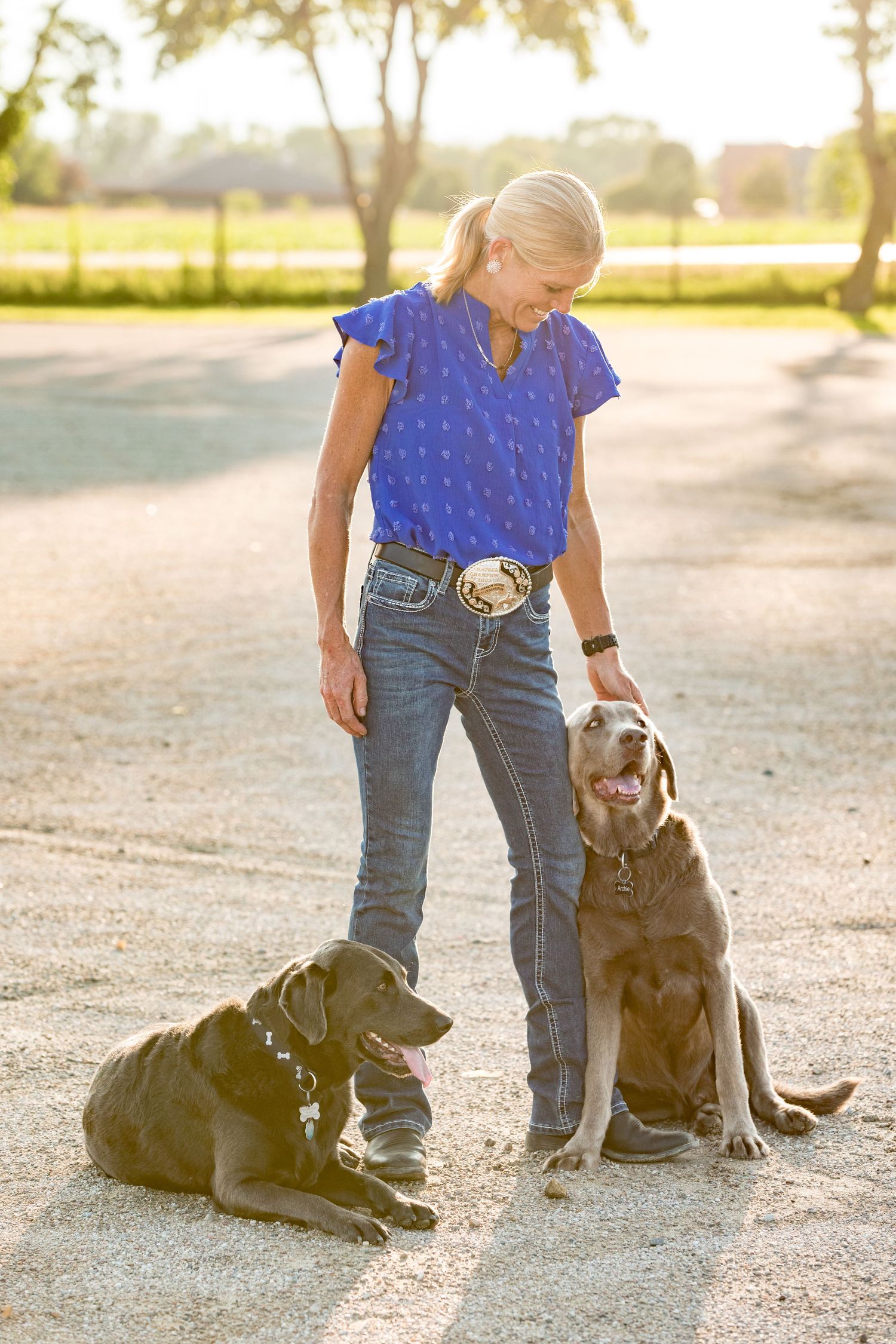 Two black Labrador dogs sitting beside someone in blue shirt and jeans on dirt path.