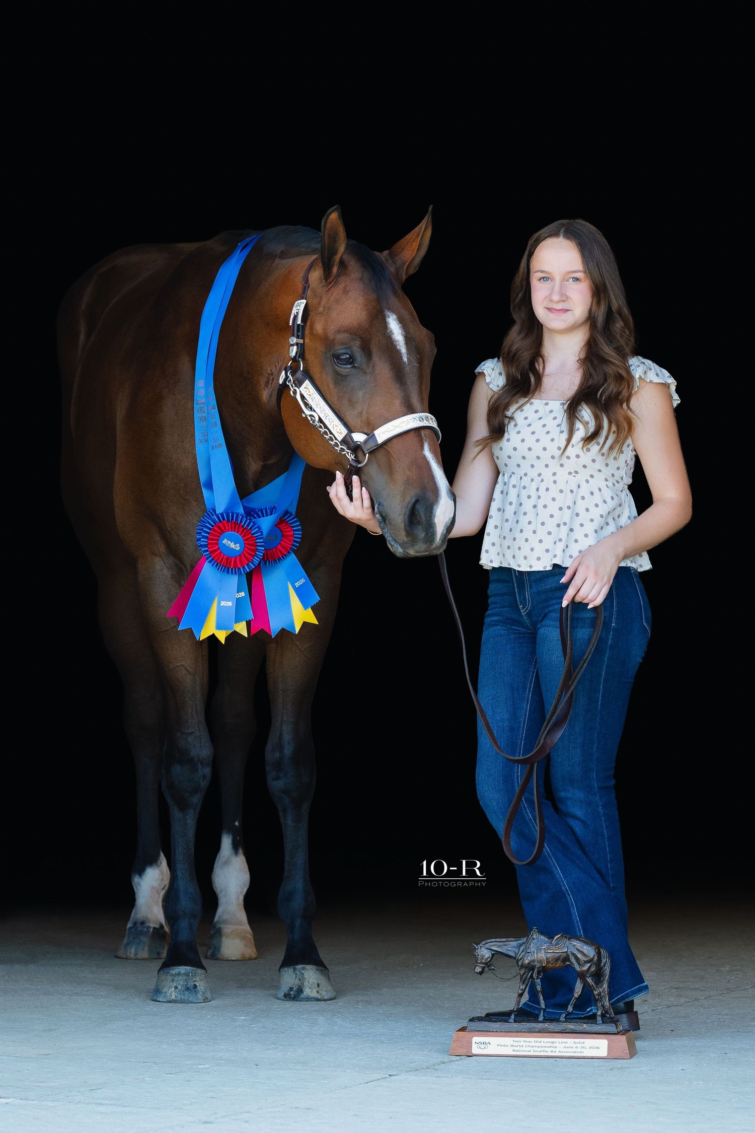 Young woman proudly poses with award-winning bay horse displaying blue and red ribbons and trophy at equestrian show.