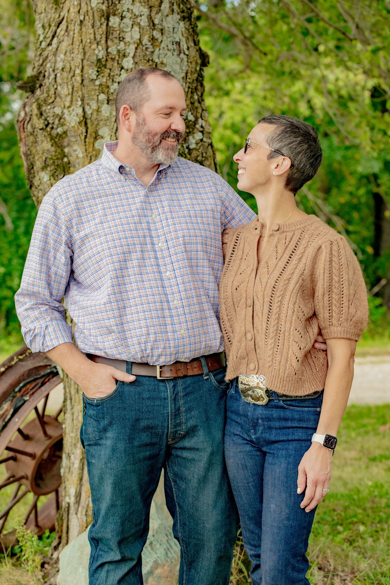 A couple shares a tender moment while standing next to a large tree trunk in a park setting.