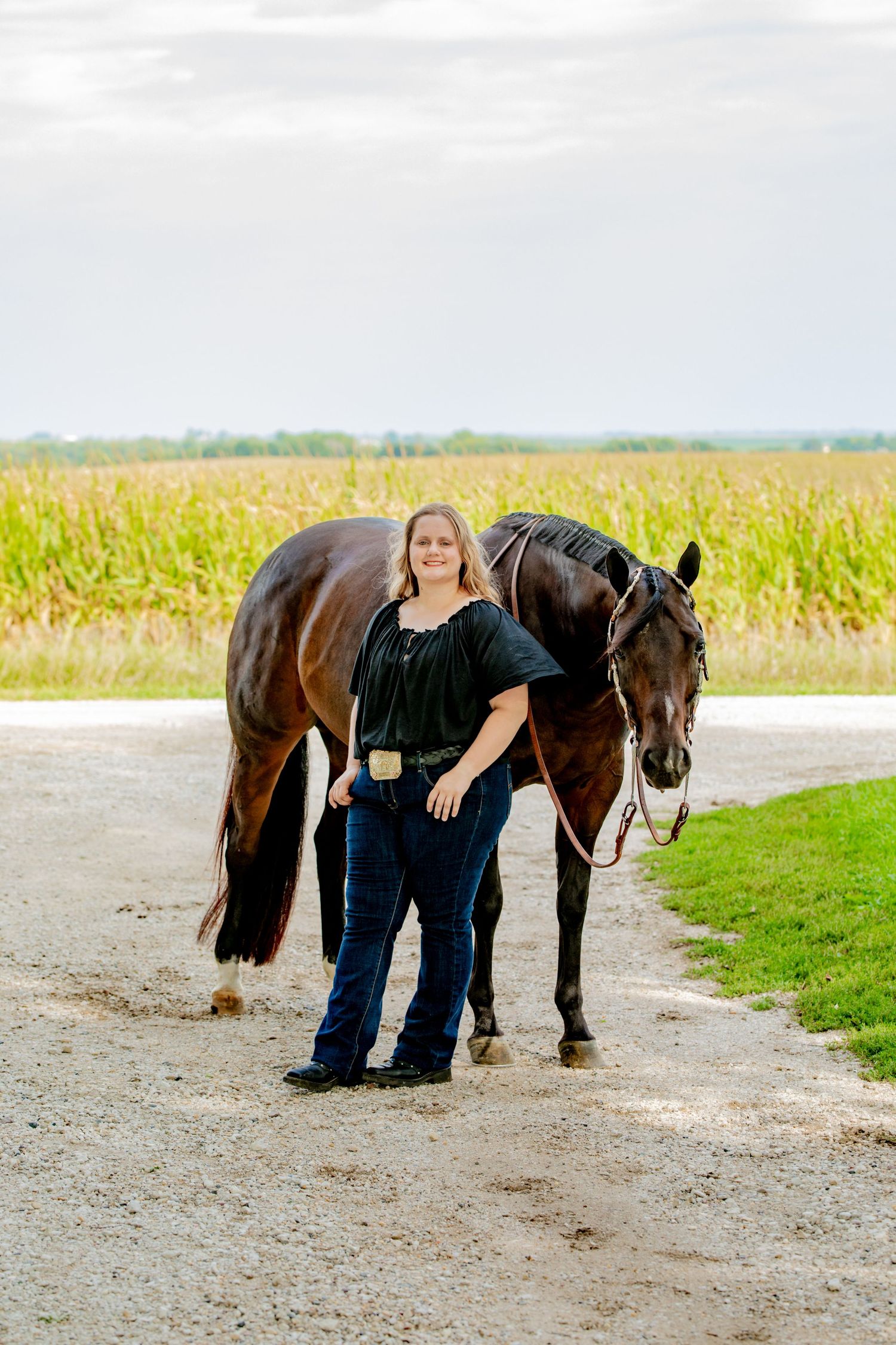 Woman in black top and jeans stands proudly beside a dark brown horse on a gravel path with cornfields behind.