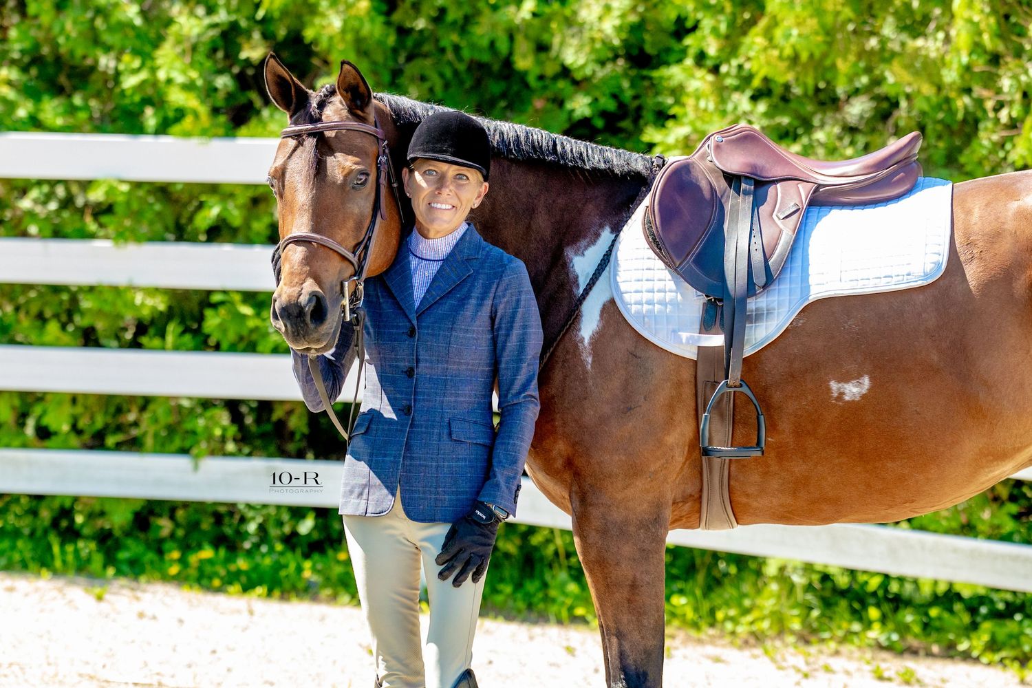 Young equestrian in blue blazer stands beside a chestnut horse with saddle at an outdoor riding arena.
