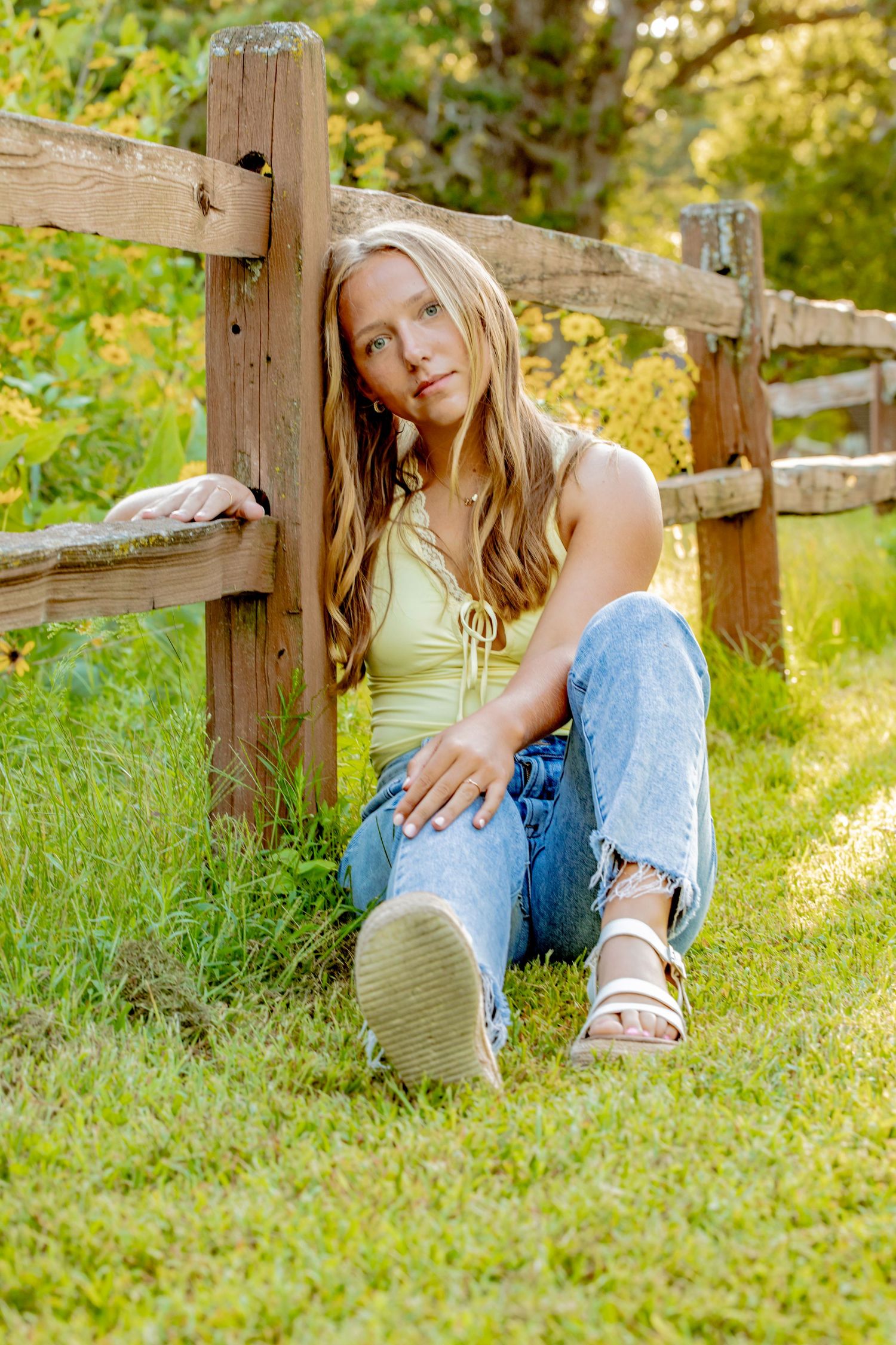 Person sitting casually by rustic wooden fence in summer sunlight wearing yellow top and jeans.