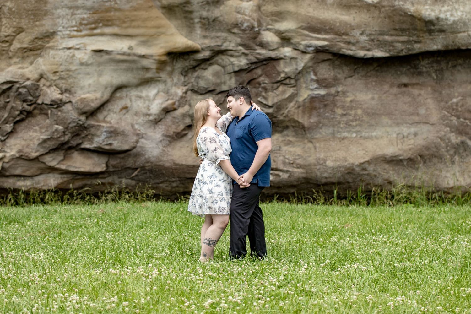 A couple embracing on green grass with rocky cliff backdrop.