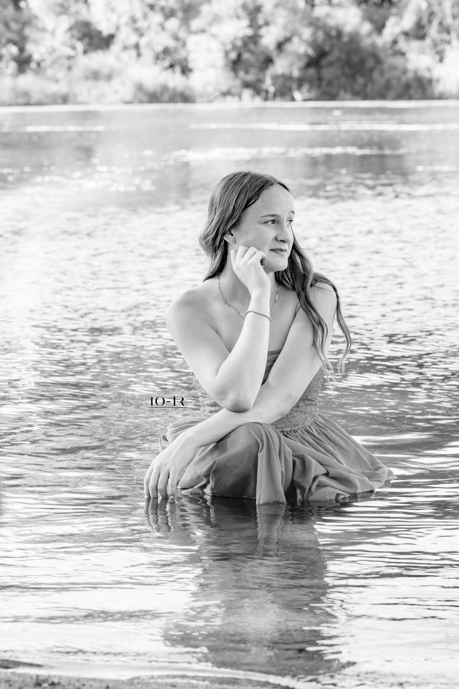 Black and white photo of a young woman sitting gracefully in a shallow river, posing thoughtfully with hand near face.