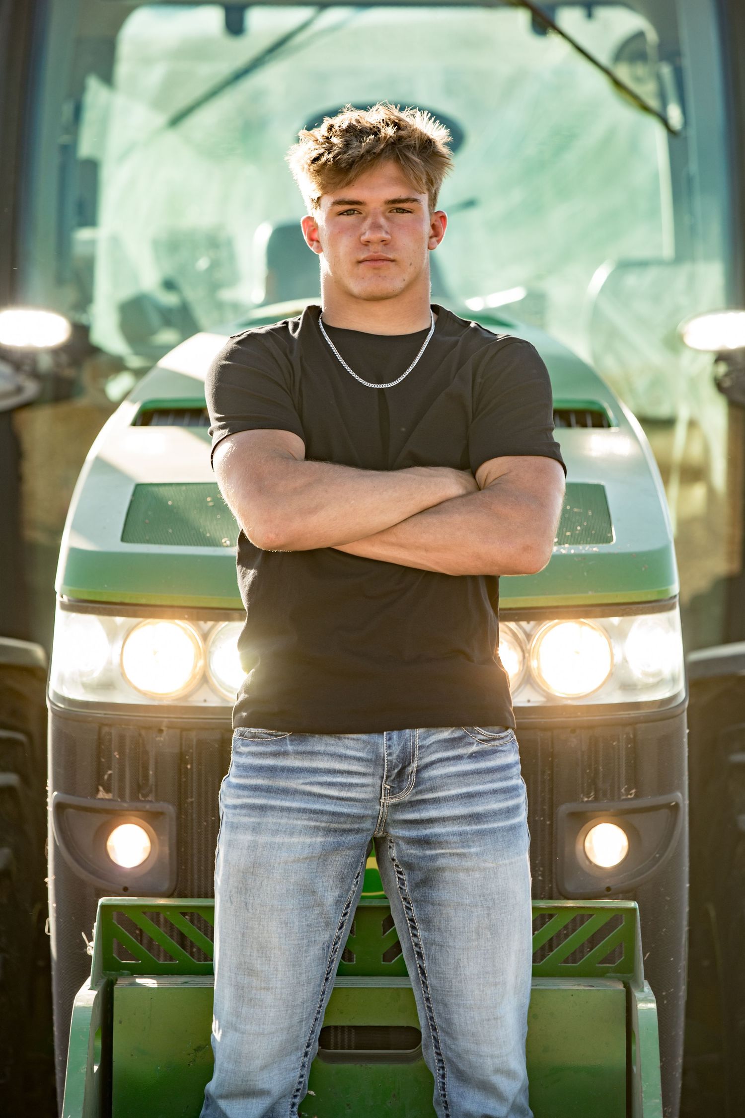 Athletic figure in black t-shirt stands with crossed arms in front of green tractor.