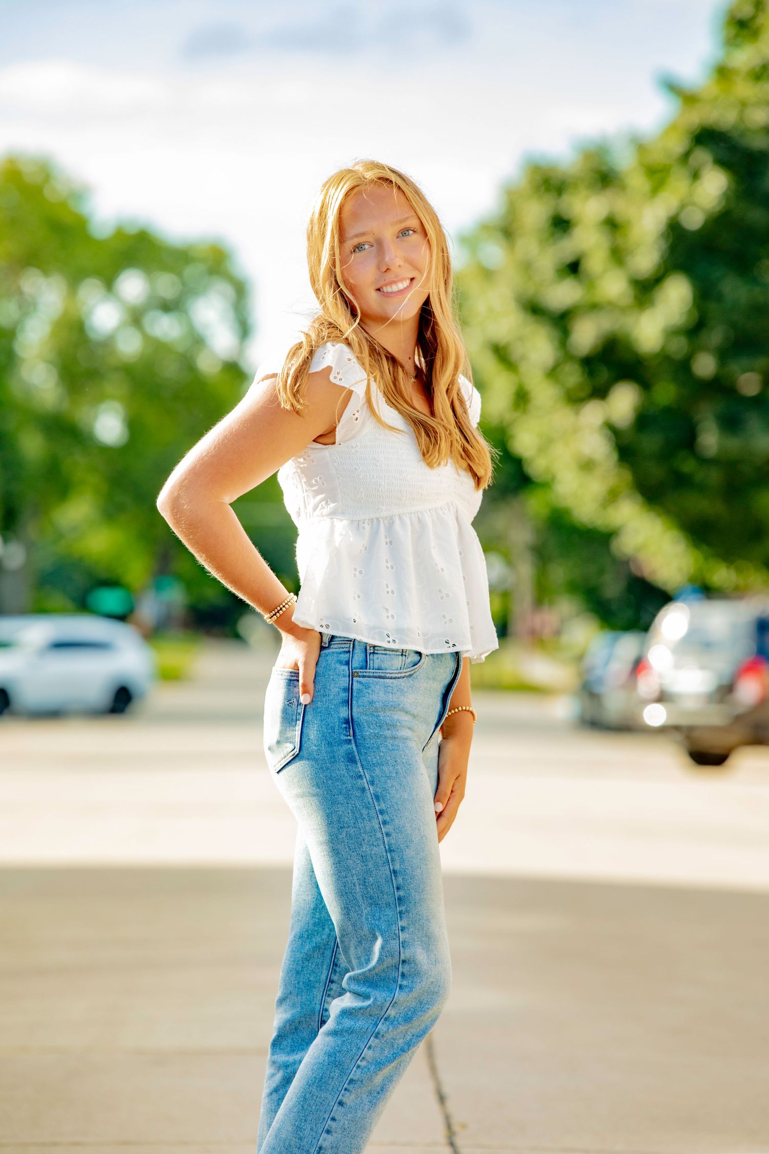 Woman in white summer top and blue jeans poses playfully outdoors in sunlit parking area.