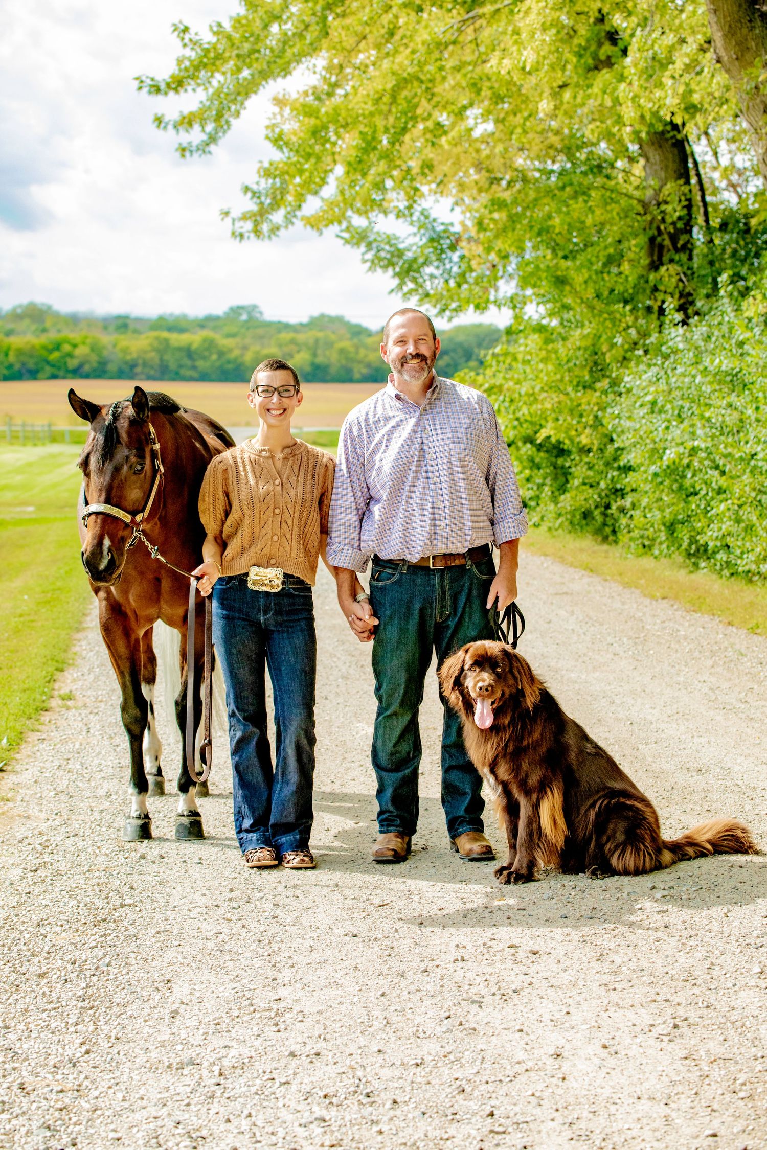 A couple and their Newfoundland dog pose on a gravel path with a horse standing behind them.